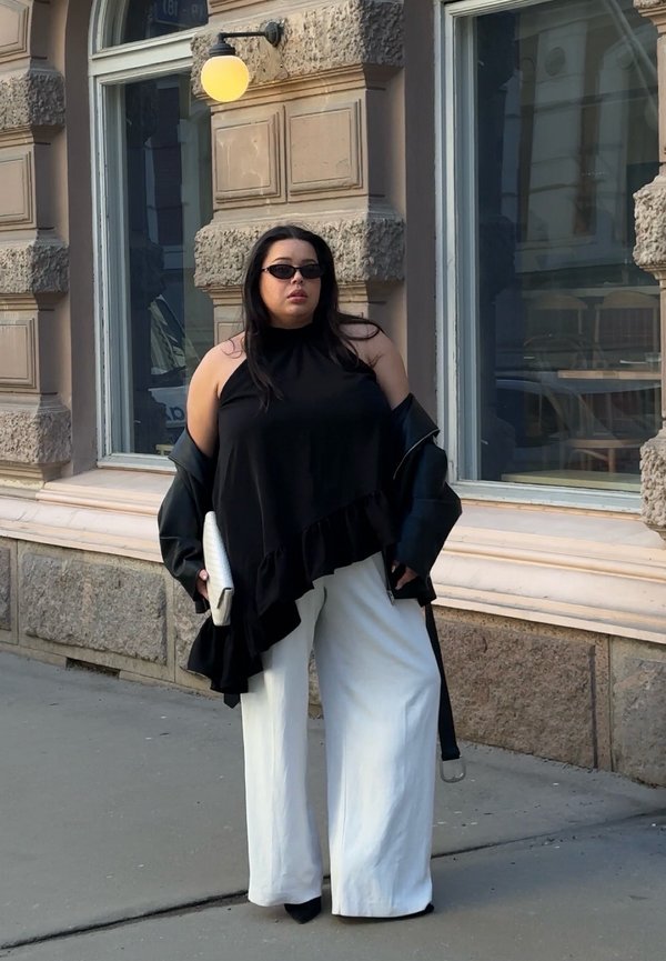 Mujer con top negro sin mangas, pantalones blancos de pierna ancha, gafas de sol, sosteniendo un bolso blanco, de pie en la acera de la ciudad.