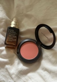 Σερum Estee Lauder Advanced Night Repair σε σκούρο γυάλινο μπουκάλι με σταγονόμετρο, δίπλα σε στρογγυλή παλέτα ρουζ MAC σε απαλό ροζ απόχρωση.