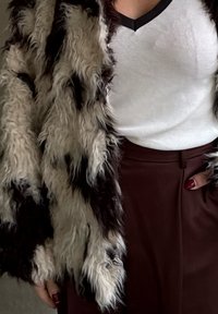 Fuskepelsjakke med en bustete tekstur i svart og krem, brukt over en hvit ribbestrikket tanktop. Brune skreddersydde bukser med sidelomme.