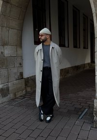 Μπεζ μακρύ παλτό, γκρι πλεκτό πουλόβερ, μαύρο παντελόνι και μαύρα-άσπρα παπούτσια. Σταθείτε σε μια καμάρα με κυβόλιο έδαφος.