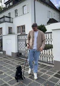 Béžový pletený kardigán, biely tričko, svetlo modré džínsy a biele tenisky. Čierny pes na vôdzke sedí vedľa osoby na vydláždenej ceste.
