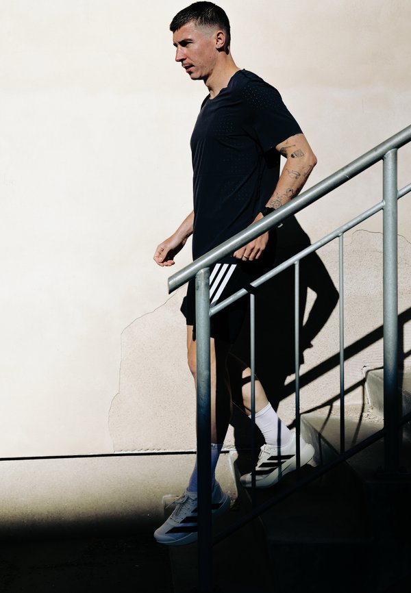 Jeune homme en tenue de sport noire descendant un escalier métallique extérieur et projetant une ombre sur un mur clair derrière lui.
