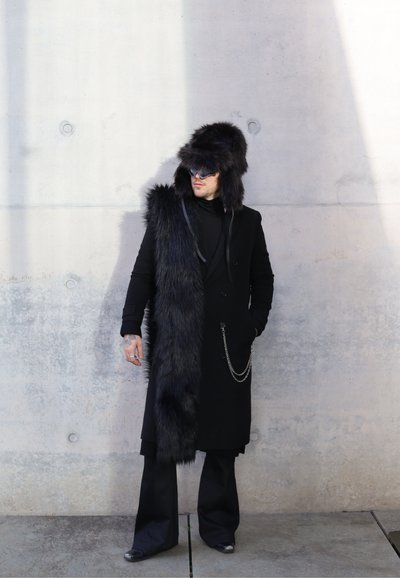 Abrigo de piel negra con acento largo y desaliñado, gorro de piel a juego y detalle de cadena plateada; combinado con pantalones negros y zapatos brillantes.