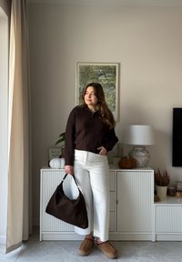 Brązowy sweter z prążkowanym wzorem, białe spodnie o prostych nogawkach, ciemnobrązowa torebka z teksturą oraz beżowe wsuwane buty. Wnętrze z elementami dekoracyjnymi.