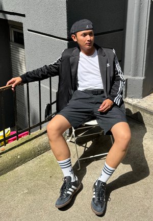 Chaqueta negra con rayas blancas, camiseta blanca, pantalones cortos negros y zapatillas negras. Calcetines blancos con rayas azules. Sentado en una silla de metal al aire libre.