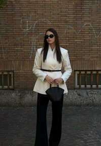 Sacou alb croitorit cu talie ajustată, pantaloni largi negri, geantă neagră și ochelari de soare rotunzi. Fundal de perete texturat din cărămidă.