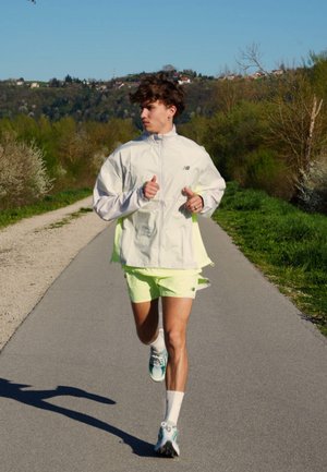 Joven con chaqueta blanca y pantalones cortos de neón corriendo por un camino pavimentado rodeado de árboles verdes y colinas bajo un cielo despejado.