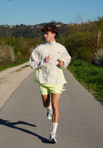 Joven con chaqueta blanca y pantalones cortos de neón corriendo por un camino pavimentado rodeado de árboles verdes y colinas bajo un cielo despejado.