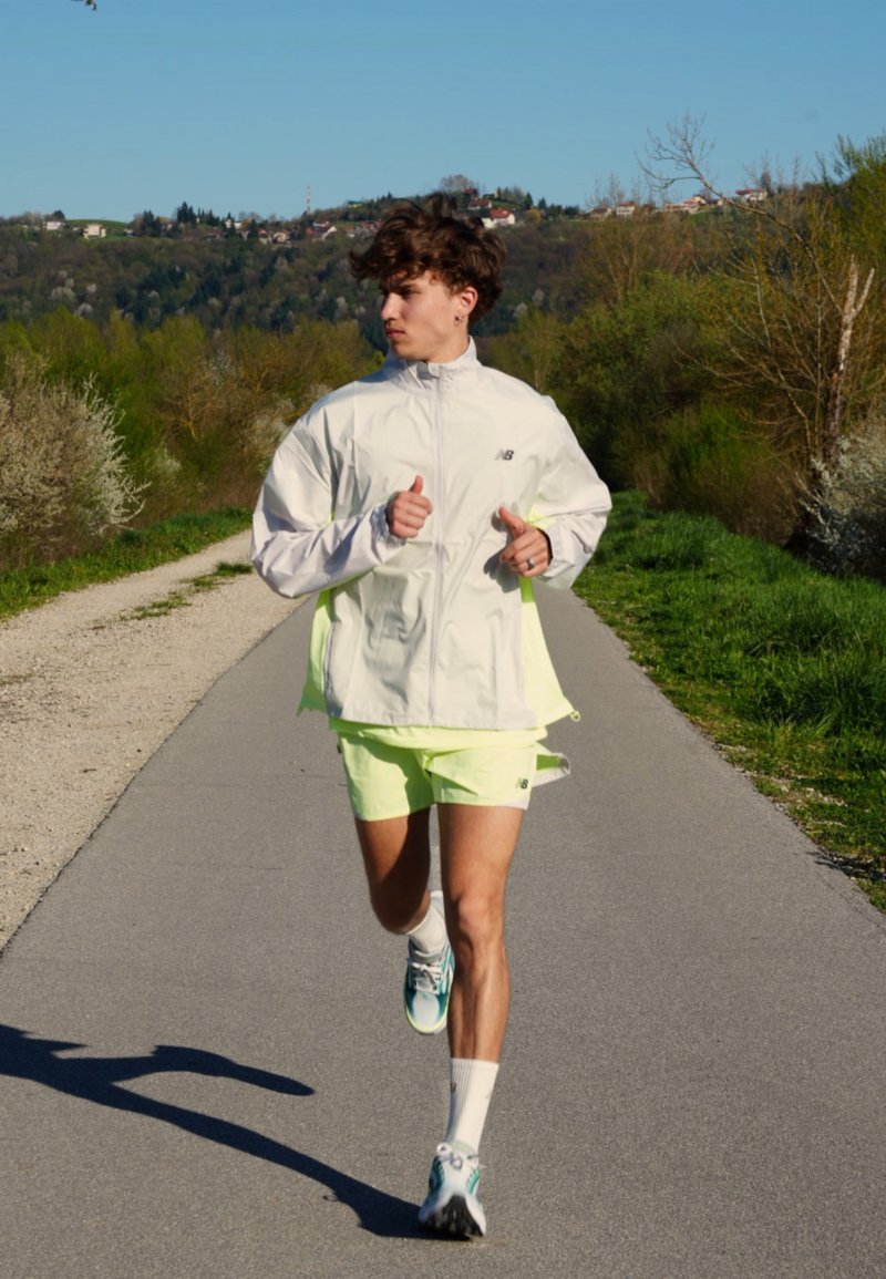 Jeune homme en veste blanche et short néon joggant sur un chemin pavé entouré d'arbres verts et de collines sous un ciel dégagé.