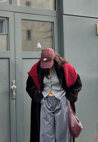 Čierny a červený nadrozmerný kabát s textúrovanými rukávmi, sivý košeľový top so zapínaním na gombíky, široké sivé nohavice a bordová čiapka, doplnené hnedou kabelkou.