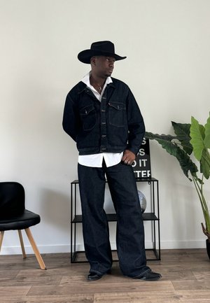 Chaqueta de mezclilla oscura con bolsillos frontales, camisa blanca de botones debajo, pantalones de mezclilla oscura a juego, sombrero de vaquero negro y botas de corte relajado.