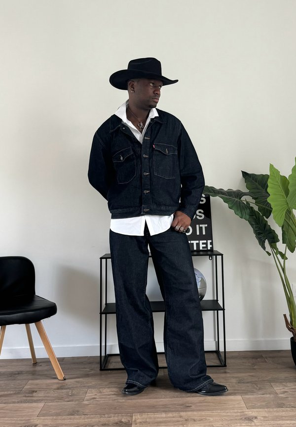 Veste en denim foncé avec poches avant, chemise blanche en dessous, pantalon en denim foncé assorti, chapeau de cow-boy noir et bottes dans une coupe décontractée.
