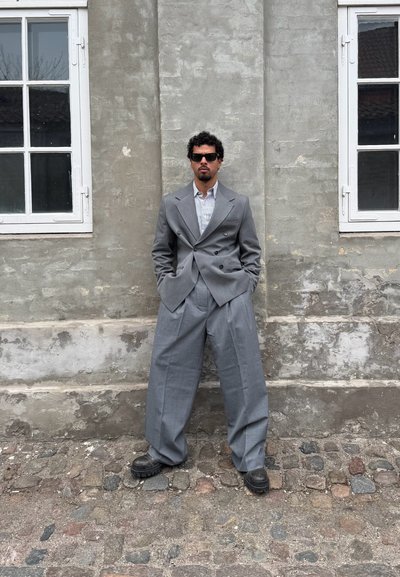 Traje gris de doble botonadura con pantalones anchos y fluidos. Combinado con una camisa de rayas, gafas de sol negras y zapatos negros gruesos contra una pared texturizada.