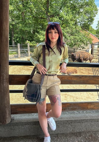 Camisa de satén verde claro, pantalones cortos beige, zapatillas blancas. Bolsa de hombro marrón con acentos de cuadros. Escenario al aire libre con animales visibles de fondo.