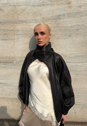 Mujer rubia con el cabello liso, vestida con una chaqueta de cuero negra sobre una blusa blanca con encaje, parada frente a una pared de piedra texturizada.