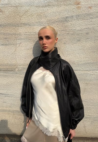 Mujer rubia con el cabello liso, vestida con una chaqueta de cuero negra sobre una blusa blanca con encaje, parada frente a una pared de piedra texturizada.