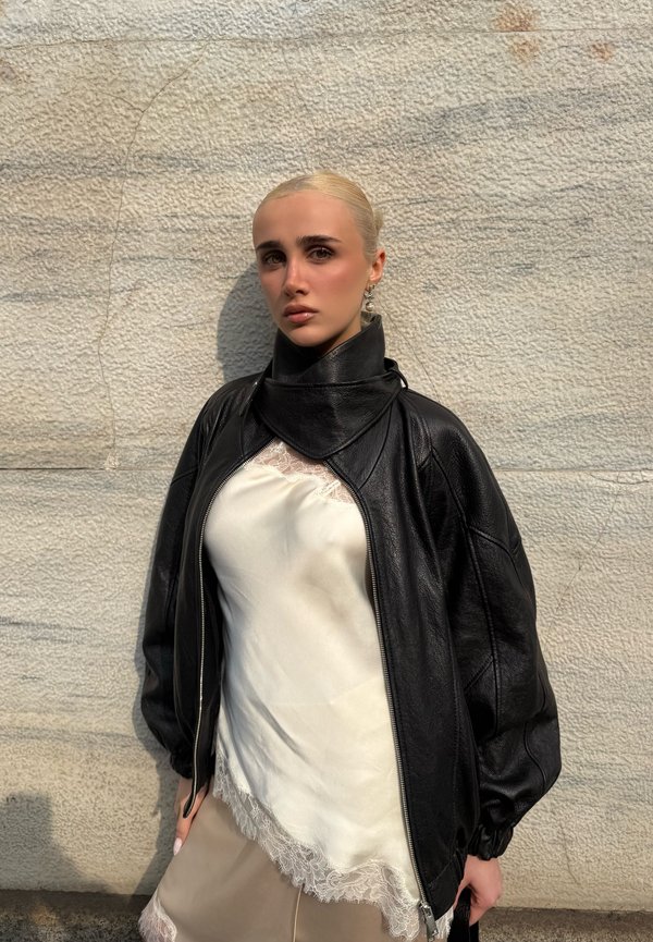 Vaaleahiuksinen nainen, jolla on sileä tukka ja joka pukeutuu mustaan nahkatakkiin valkoisen pitsireunaisen topin päällä, seisoo teksturoidun kiviseinän edessä.