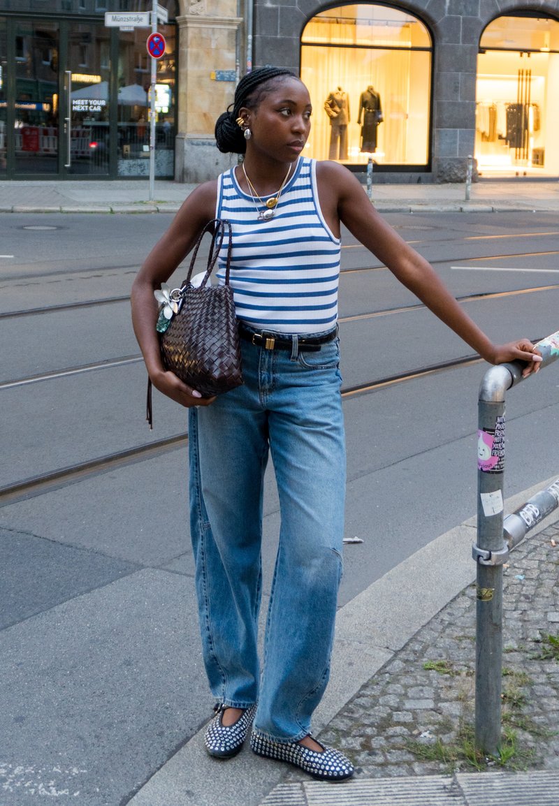 Young woman in a striped tank top, blue jeans, and studded shoes stands on a city sidewalk, holding a dark woven bag and resting her hand on a metal railing.