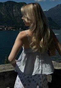 Top sem mangas azul claro com cintura franzida; cabelo longo e ondulado loiro; saia branca com pequenos padrões florais; fundo de paisagem natural.