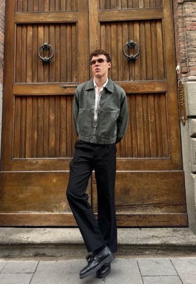 Chaqueta corta verde oliva sobre una camisa blanca, combinada con pantalones de pierna ancha negros y zapatos negros, con un fondo de puerta de madera.