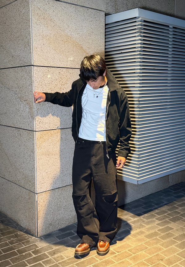 Jeune homme portant une veste noire, une chemise blanche et un pantalon noir, debout contre un mur en angle avec une ventilation industrielle dans un cadre urbain la nuit.