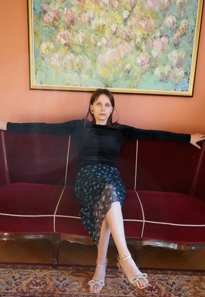 Sofá de terciopelo rojo con detalles dorados, blusa de manga larga negra, falda azul con estampado y sandalias ligeras de tiras. Pintura floral en el fondo.