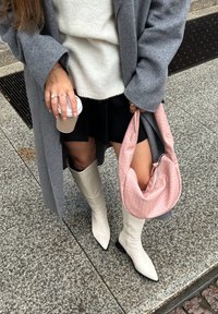 Grau oversized Mantel, weißer Strickpullover, schwarzer Rock, weiße spitze Stiefel und eine rosa strukturierte Tasche. Hält eine Kaffeetasse.