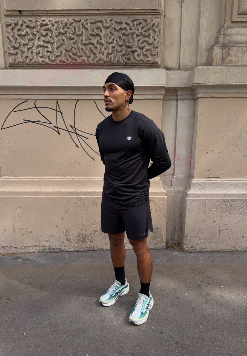 Homme portant des vêtements de sport noirs et des baskets bleu sarcelle debout, les mains dans le dos, contre un mur en pierre couvert de graffitis.