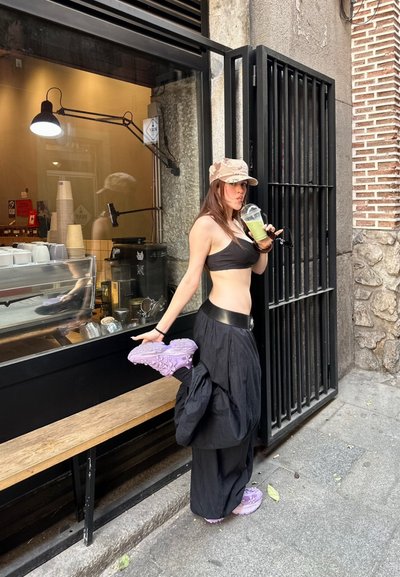 Top corto negro y falda negra fluida, zapatos morados texturizados, gorra beige estampada, sosteniendo una bebida afuera de un café con banco de madera.