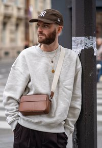 Brun lær crossover veske med en flat frontlomme, justerbar stropp og kontraststing, brukt av en person i en grå hettegenser.