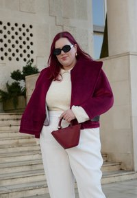 Bordó színű velúr dzseki széles hajtókkal, krémszínű pulóver és krémszínű nadrág. Egy kicsi, vörös, strukturált kialakítású kézitáskát tart.