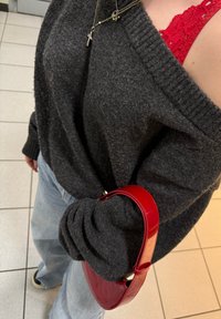 Donkergrijze oversized trui met geribbelde manchetten, gecombineerd met lichtblauwe jeans. Houdt een glanzende rode handtas met een ronde handgreep vast.