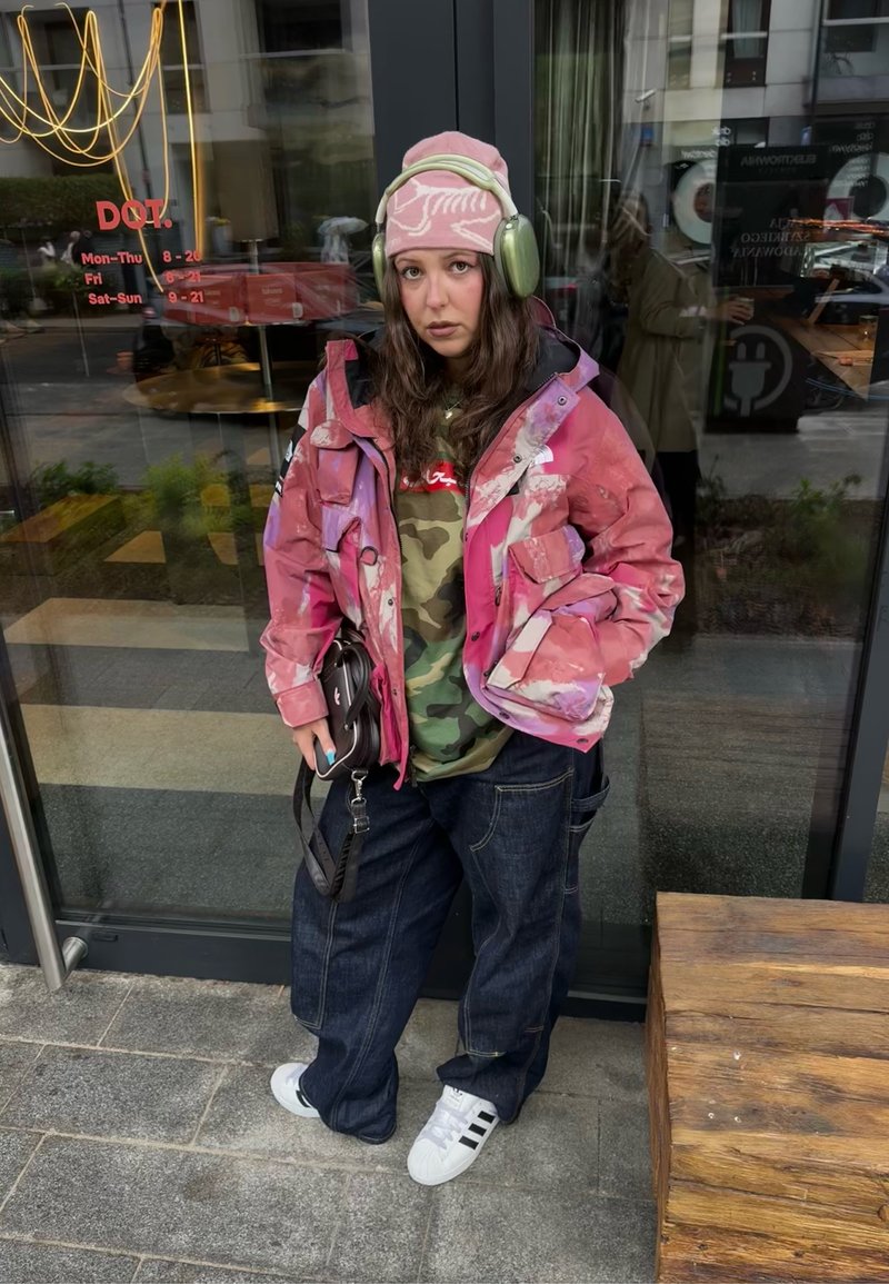Jeune personne portant un bonnet rose et une veste, des écouteurs verts, un t-shirt camouflage, un jean large foncé et des baskets blanches, debout devant une porte vitrée.