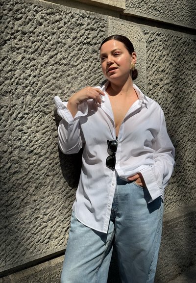 Mujer con cabello oscuro recogido hacia atrás se apoya en una pared de piedra texturizada, lleva una camisa blanca con botones, vaqueros azules y gafas de sol sujetas a la camisa.
