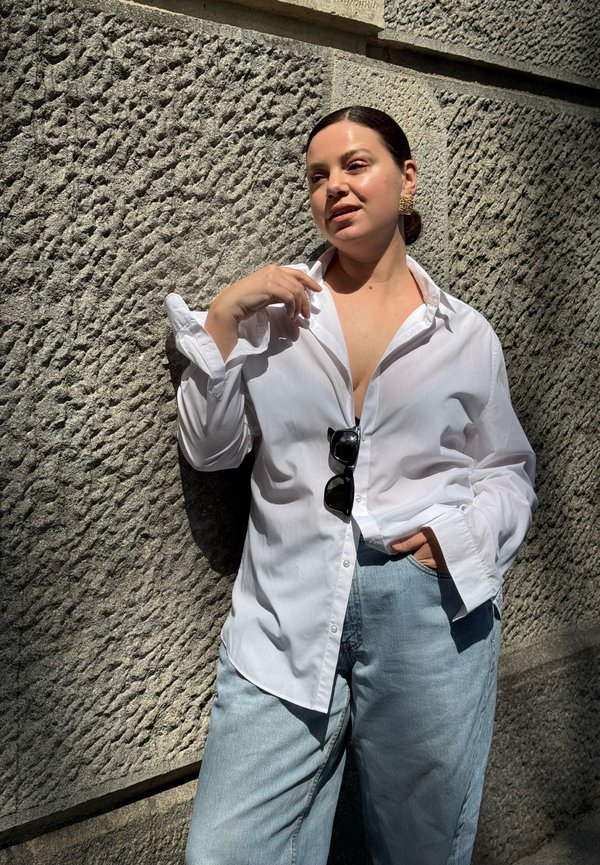 Vrouw met donker haar naar achteren gebonden leunt tegen een getextureerde stenen muur, draagt een wit overhemd met knopen, blauwe spijkerbroek en een zonnebril vastgeclipt aan het overhemd.