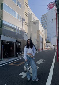 Μοντέλο σε λευκό cropped top και ανοιχτού μπλε φλαρέ παντελόνι, κρατώντας μια τσάντα με κεντήματα, με μια γκρ�ι φούτερ ριχτή στους ώμους της.