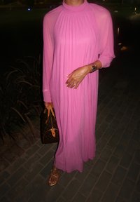 Lang, flytende rosa kjole med plisserte detaljer, mykt stoff og høy hals. Gyldne sandaler med stropper og en brun håndveske med logo.