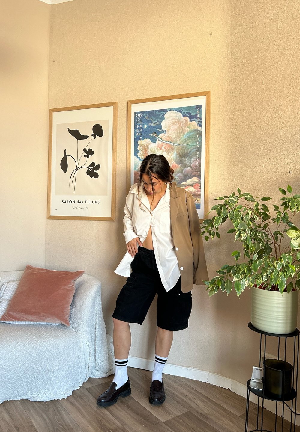 Καθημερινό ντύσιμο με μπεζ σακάκι, λευκό πουκάμισο, μαύρες βερμούδες, λευκές κάλτσες με μαύρες ρίγες και μαύρες loafers. Εσωτερικός χώρος με διακοσμητική τέχνη.