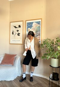 Καθημερινό ντύσιμο με μπεζ σακάκι, λευκό πουκάμισο, μαύρες βερμούδες, λευκές κάλτσες με μαύρες ρίγες και μαύρες loafers. Εσωτερικός χώρος με διακοσμητική τέχνη.