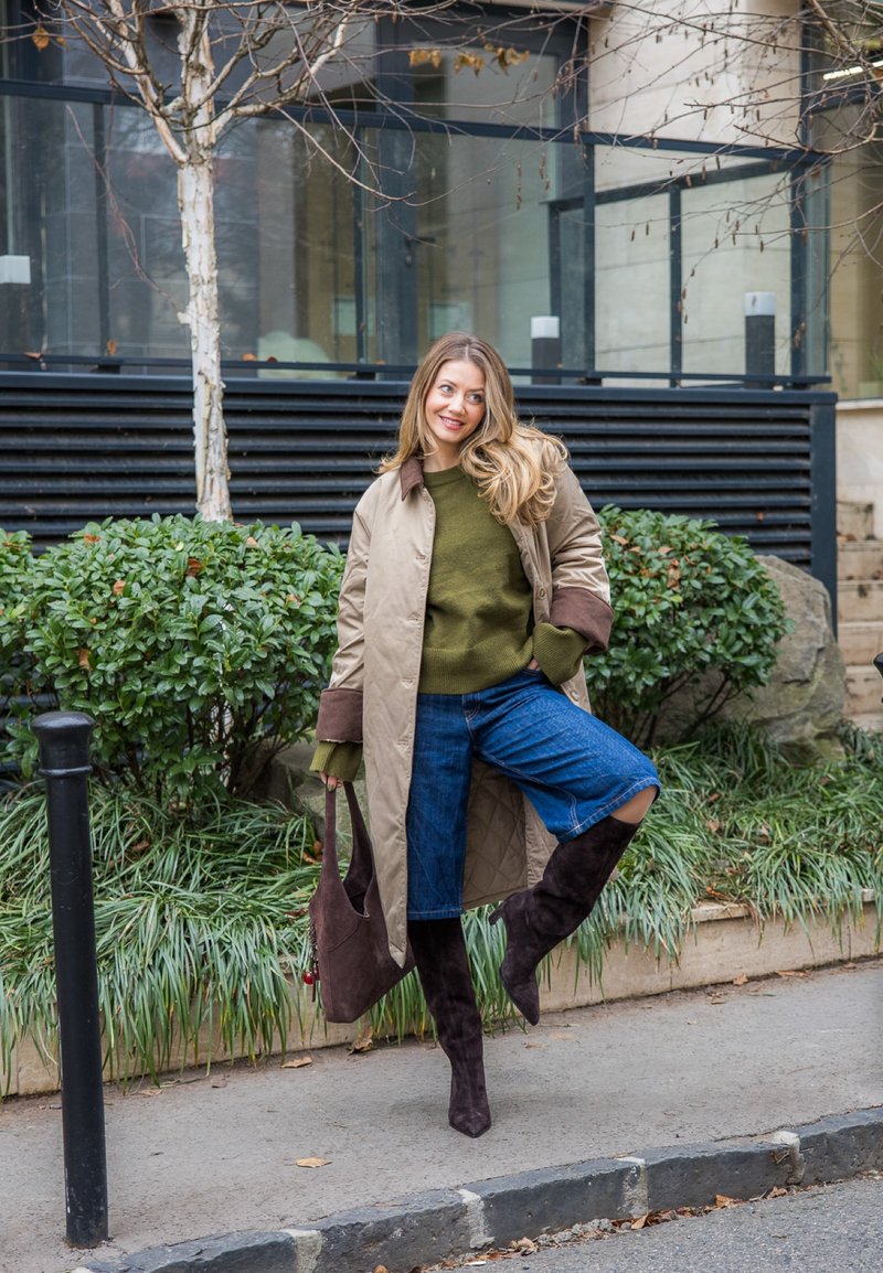 Femme en manteau beige, pull vert, shorts en denim et bottes montantes posant sur le trottoir avec une jambe levée, portant un sac marron en extérieur.