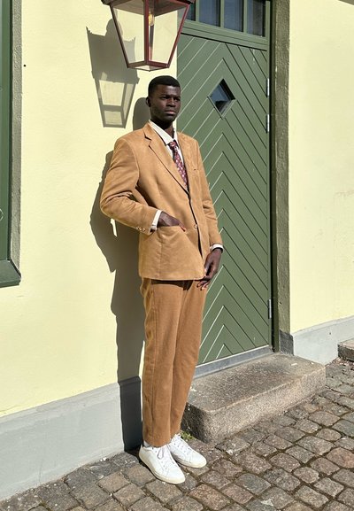 Hombre con traje color beige y corbata estampada apoya contra una pared amarillo claro con una puerta verde y un farol, de pie sobre un pavimento de adoquines.