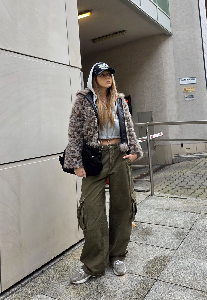 Syntetická kožená bunda s kapucí, šedý crop top, olivově zelené cargo kalhoty a šedé tenisky. Černá kšiltovka a kabelka dokončují vzhled.