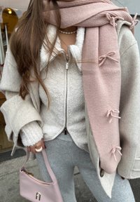 Femme portant une veste en polaire crème, un manteau beige, une écharpe rose avec des nœuds, un pantalon gris, un collier de perles, et tenant un sac à main rose.