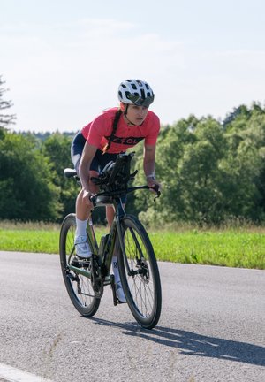 Atleta su una elegante bicicletta da strada nera, indossando una maglia rosa brillante e pantaloni corti scuri, in una postura concentrata, con casco bianco e scarpe da ciclismo.