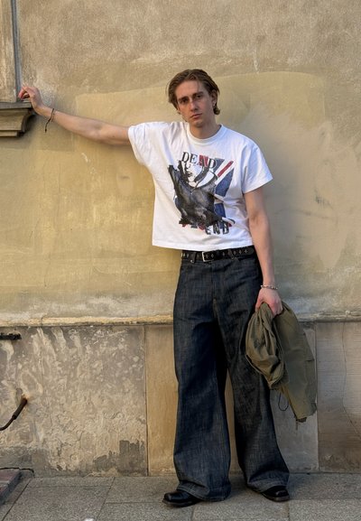 Joven apoyado contra una pared beige, con camiseta blanca con diseño, vaqueros anchos oscuros, sosteniendo una chaqueta verde en la mano, mirando a la cámara.