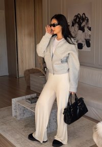 Cápsula cinza com fecho, camiseta branca, calças largas de creme, óculos de sol pretos e uma mala preta. Texturas suaves e design casual.