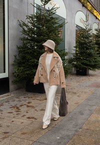 Béžový kabát dvojradový z ​​vlneného zmesi s kvetinovou výšivkou, krémový sveter, biele nohavice, sivá taška na rameno a béžové lodičky. V pozadí borovicové stromy.