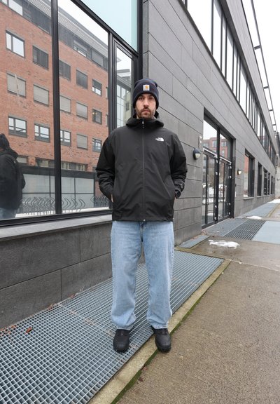 Hombre con chaqueta negra, gorro azul marino, vaqueros azul claro y zapatos negros de pie en la acera junto a un edificio moderno con ventanas de cristal.