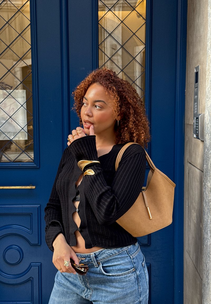 Μαύρο ριγέ κασκόλ με ανοιχτά κουμπιά, ανοιχτό καφέ γωνιακό τσαντάκι, τζιν παντελόνι ψηλής μέσης και χρυσά αξεσουάρ μπροστά από μια μπλε πόρτα.