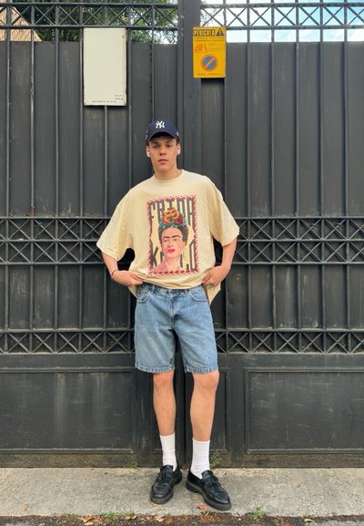 Camiseta beige de gran tamaño con estampado de Frida Kahlo, pantalones cortos de mezclilla, zapatillas negras y calcetines blancos, de pie frente a una puerta de metal gris.
